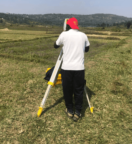 Slide 2 &mdash; A surveyor in a red cap stands behind a leveling instrument on a tripod in a lush green field, with hills in the background.