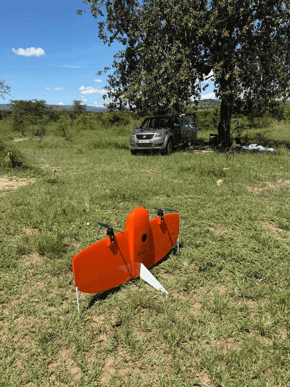Slide 5 &mdash; An orange drone rests on grassy terrain beside a parked silver SUV under a tree, with blue skies and distant hills in the background.