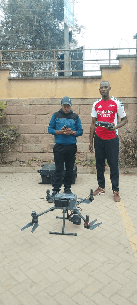 Slide 6 &mdash; Two individuals stand beside a drone on a paved surface, one checking a device while the other holds a controller, with a wall in the background.