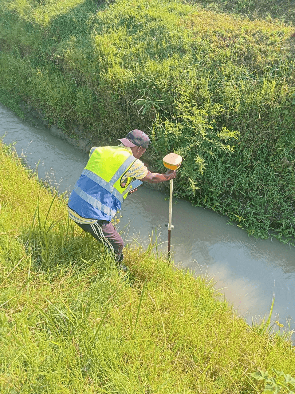 Slide 7 &mdash; A worker in a high-visibility vest uses surveying equipment near the edge of a grassy drainage canal.